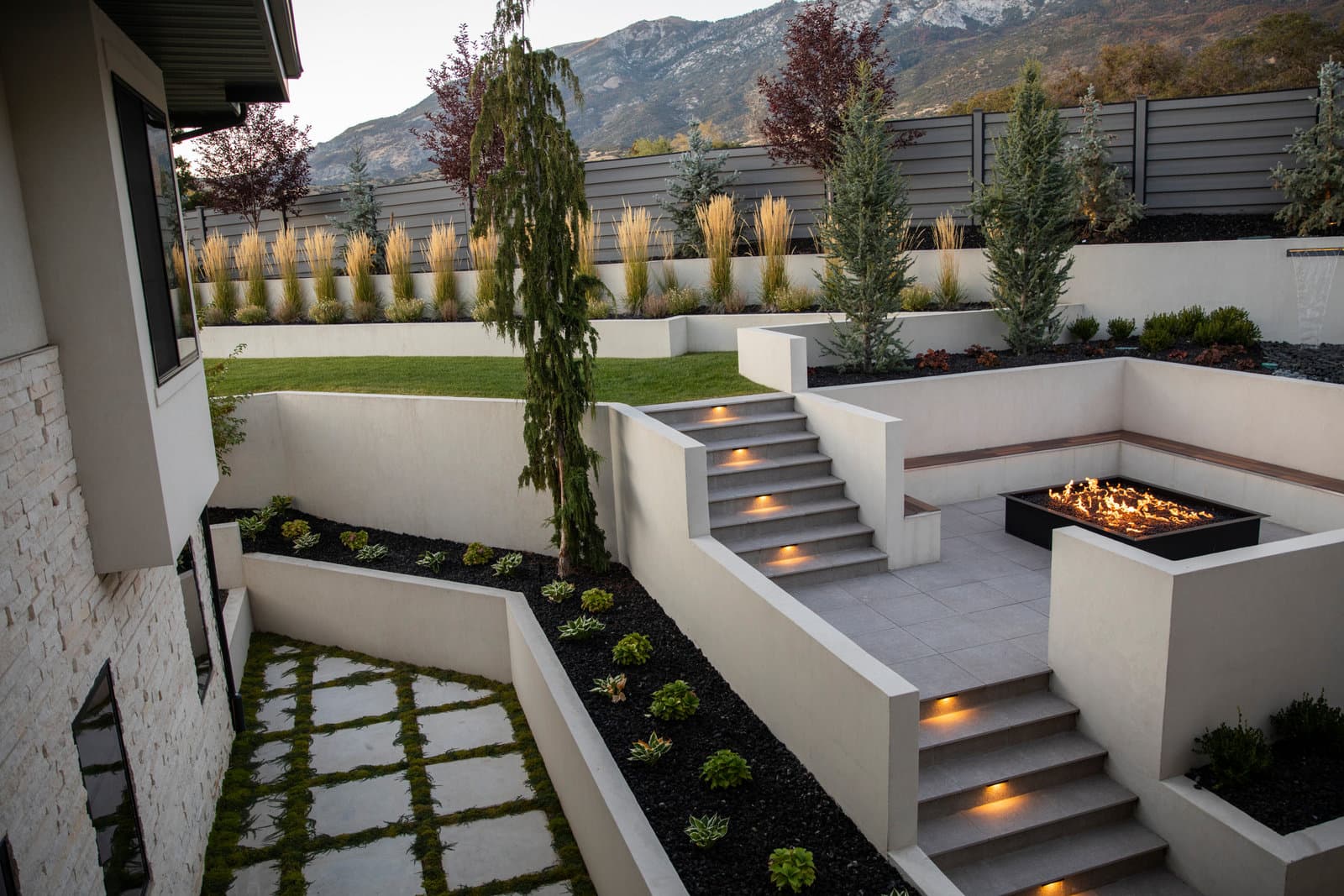 Big Rock modern sunken courtyard with steel fire pit, lit stone stairs, raised planters, and Wasatch mountain backdrop in Alpine, Utah