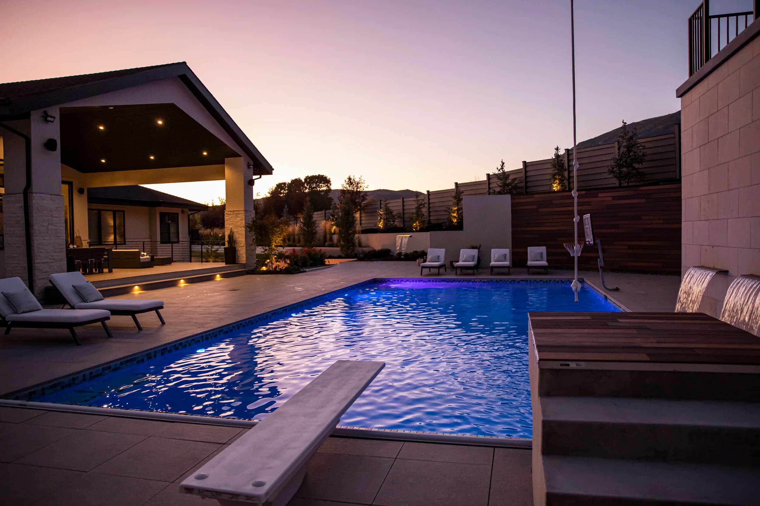 Big Rock custom pool with diving board, illuminated water, covered patio, and Wasatch mountain twilight at the Frank residence in Alpine, Utah