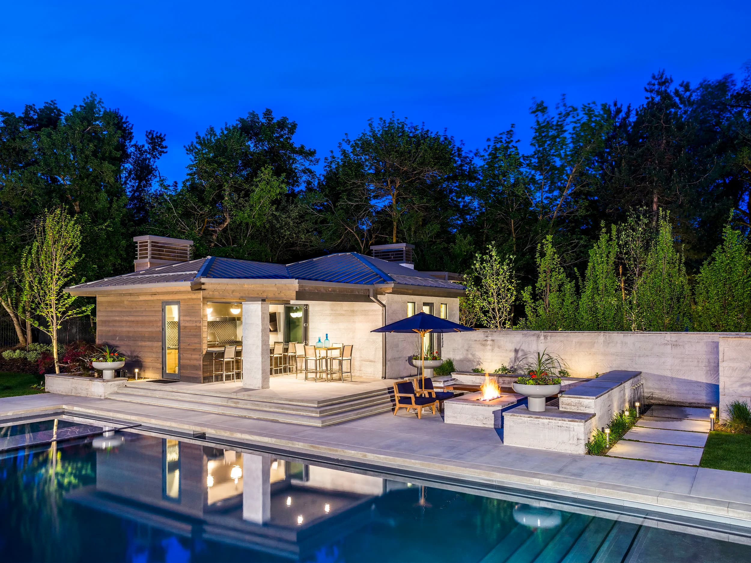 Contemporary Big Rock poolhouse mirrored in a still pool at blue hour, with built-in firepit and bar seating