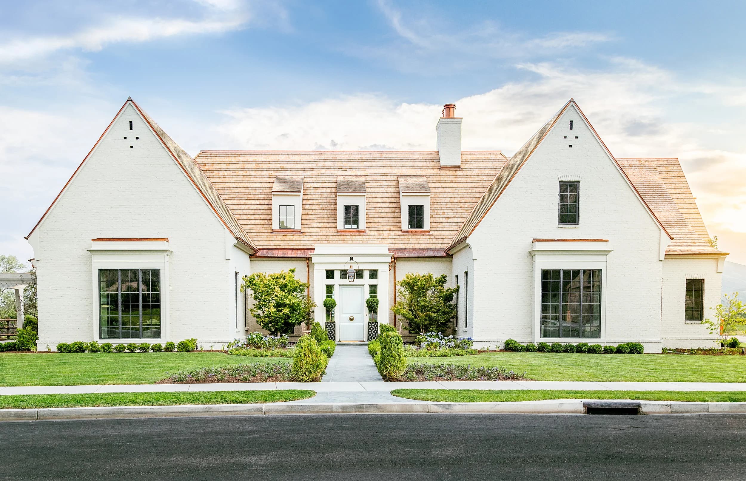 Symmetrical white Tudor estate with cedar shake roof, manicured topiary, and golden Utah sunset